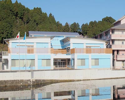水色と白を基調とした介護施設の外観。川沿いに建つ平屋の建物で、周囲に緑が豊富。